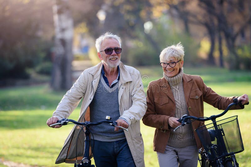 Cheerful active senior couple with bicycles walking through park together. Perfect activities for elderly people.