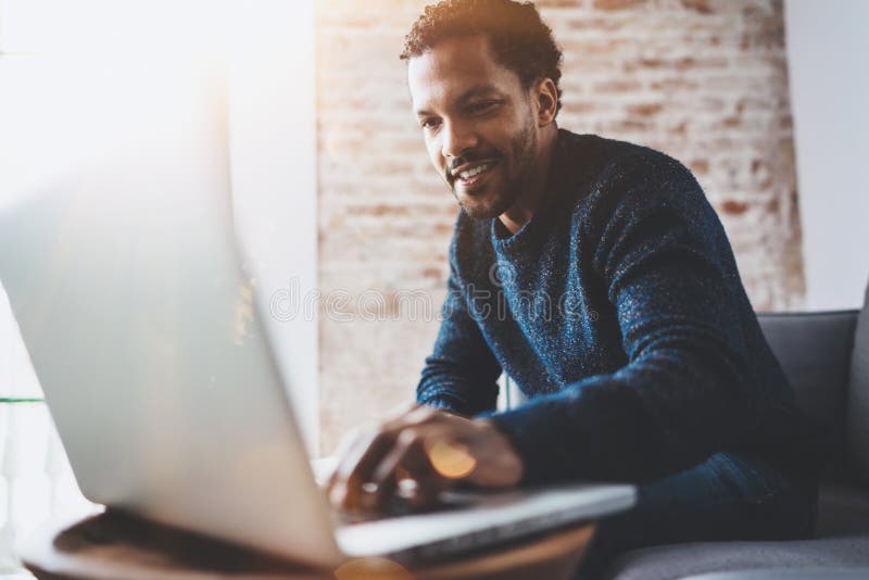Cheerful African man using computer and smiling while sitting on the sofa.Concept of young business people working at. Home.Blurred background,flares stock image
