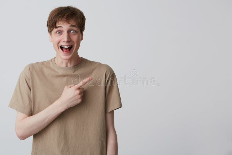 Cheerful amazed young man student with metal braces on teeth wears beige t shirt and points to the side  over white