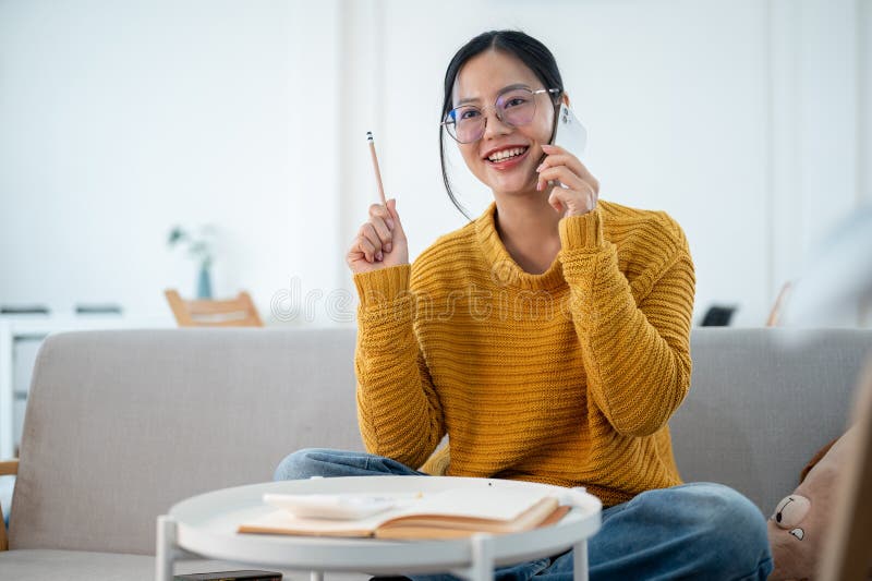 A cheerful Asian woman is sitting on a sofa, talking on the phone while making a list in a notebook royalty free stock images