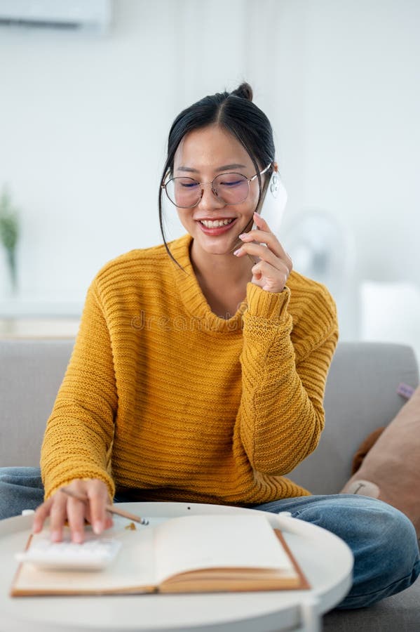 A cheerful Asian woman is sitting on a sofa, talking on the phone while making a list in a notebook stock image