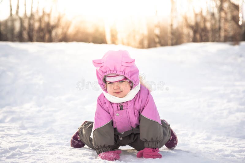 Cheerful baby on all fours playing on snow in cold sunny winter day in warm clothes with sunlight through trees stock photo