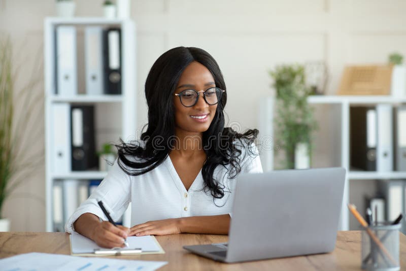 Cheerful black business lady working on laptop, taking notes during online work meeting or webinar at office