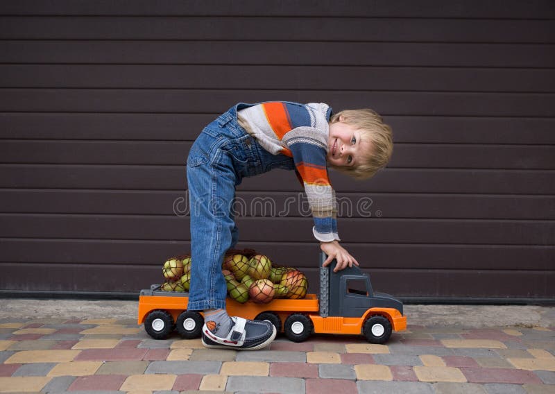 Cheerful boy 4-5 years in denim overalls and knitted sweater carries apples in string bag on toy car truck