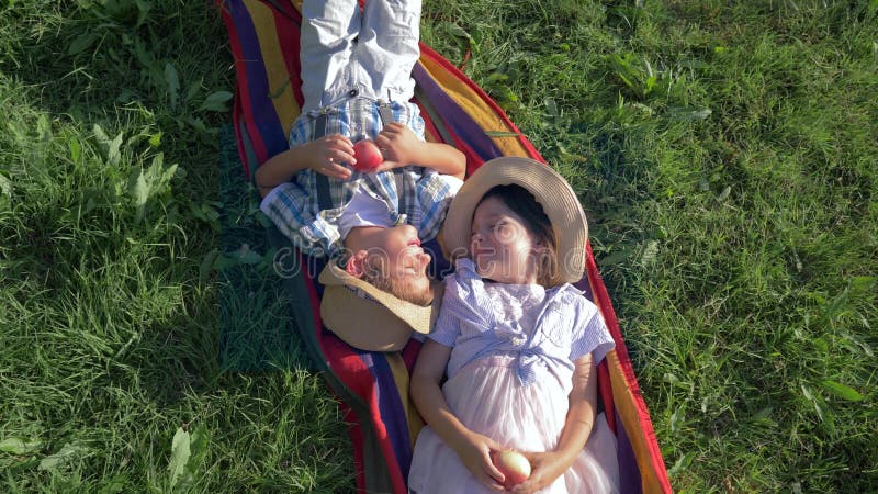 Cheerful children in straw hats are lying on blanket with apples in their hands and communicate at summertime