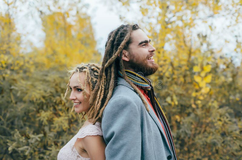 A cheerful couple stands back to back. Bride and groom with dreadlocks having fun on nature. Wedding ceremony outdoors.