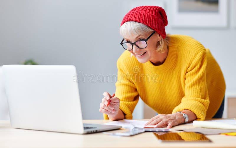 Cheerful elderly woman creative designer in red hat smiles at workplace at  table with a laptop, sketches and samples