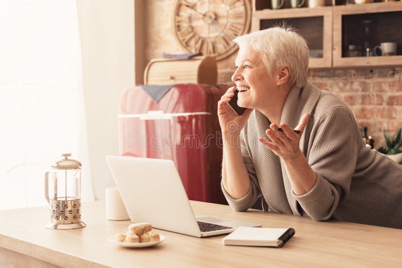 Cheerful elderly woman talking on cellphone and using laptop in kitchen