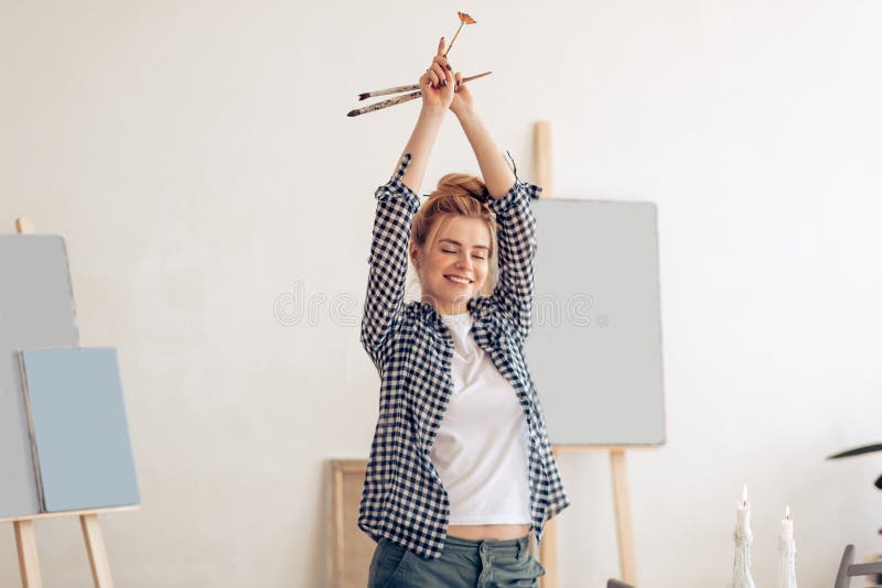 Cheerful funny girl with hairbun raising her arms and holding brushes
