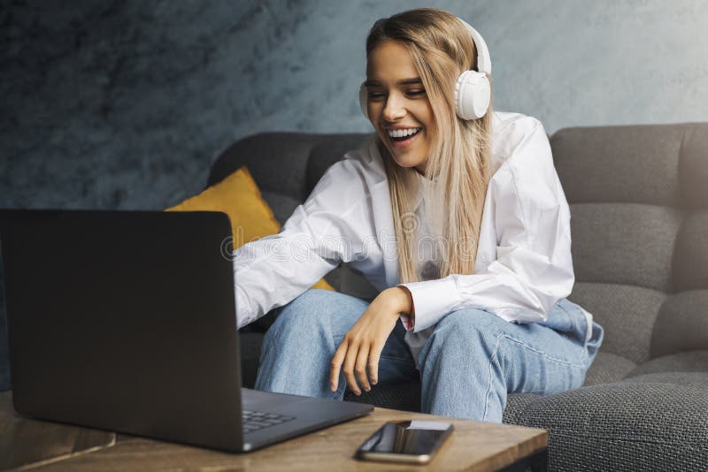 Cheerful girl in headphones sitting on sofa in front of laptop. Entertainment online content for streaming at affordable