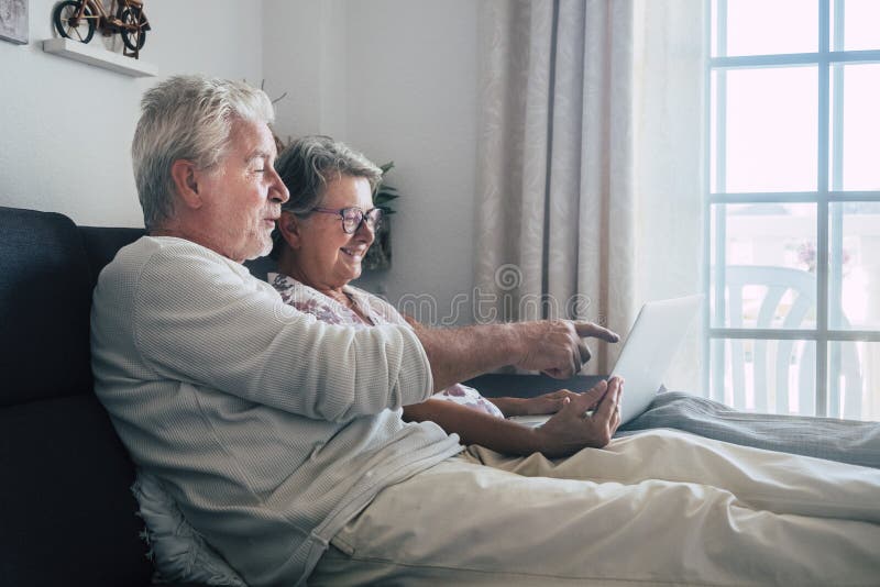 Cheerful happy beautiful caucasian old aged senior couple sit down and relax on the soa at home looking and using a personal