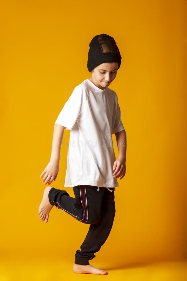 Cheerful little boy, dressed in sweatpants and a white T-shirt, actively dancing, standing on one leg. yellow background