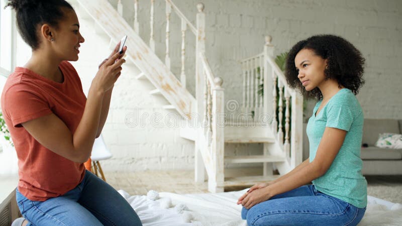 Cheerful mixed race woman photographing his girlfriend using smartphone camera sitting on bed in bedroom at home royalty free stock photography Cheerful mixed race woman photographing his girlfriend using smartphone camera sitting on bed in bedroom at home royalty free stock photography
