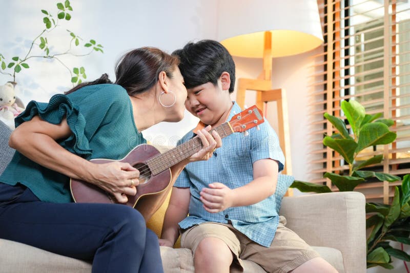 Cheerful Moder och Son Enjoying Ukulele Music Together at Home Asian Woman Teaching Kid Boy to Play Ukulele in Living Room arkivbild