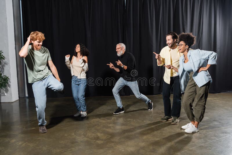 Cheerful multicultural actors pointing at redhead man rehearsing on stage in theater,stock image