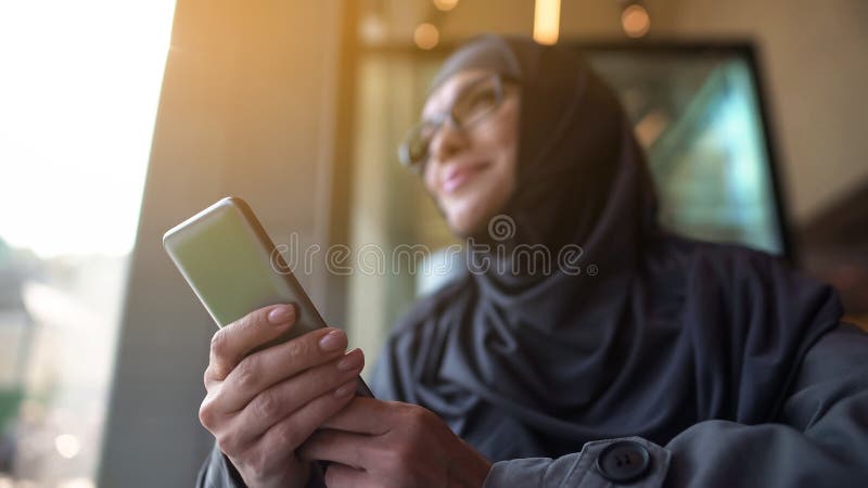 Cheerful Muslim lady holding phone in hand, looking outside through cafe window