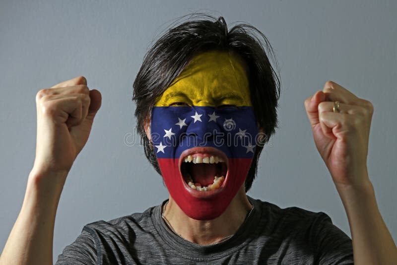 Cheerful portrait of a man with the flag of Venezuela painted on his face on grey background. The concept of sport or nationalism.