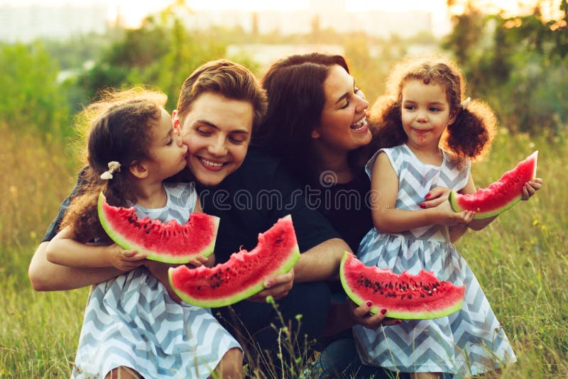 Cheerful positive family of four having a picnic and eating watermelon outdoors in a sunny weather. Curly beautiful twins sisters stock photography Cheerful positive family of four having a picnic and eating watermelon outdoors in a sunny weather. Curly beautiful twins sisters stock photography