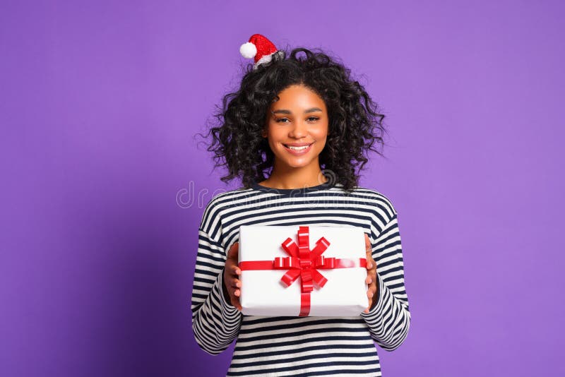 Cheerful smiled  ethnic woman  with santa hat   holds a Christmas gift on colorful  purple  background