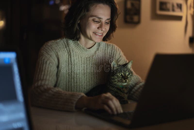 Cheerful smiling woman 30 years old sitting in front of a laptop at home in the evening with a gray pet cat in her arms