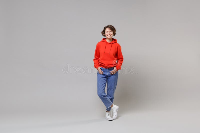 Cheerful young brunette woman girl in casual red hoodie, blue jeans posing isolated on grey background studio portrait