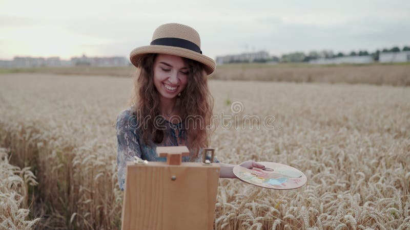 Cheerful young lady in hat painting with watercolors among ripe wheat