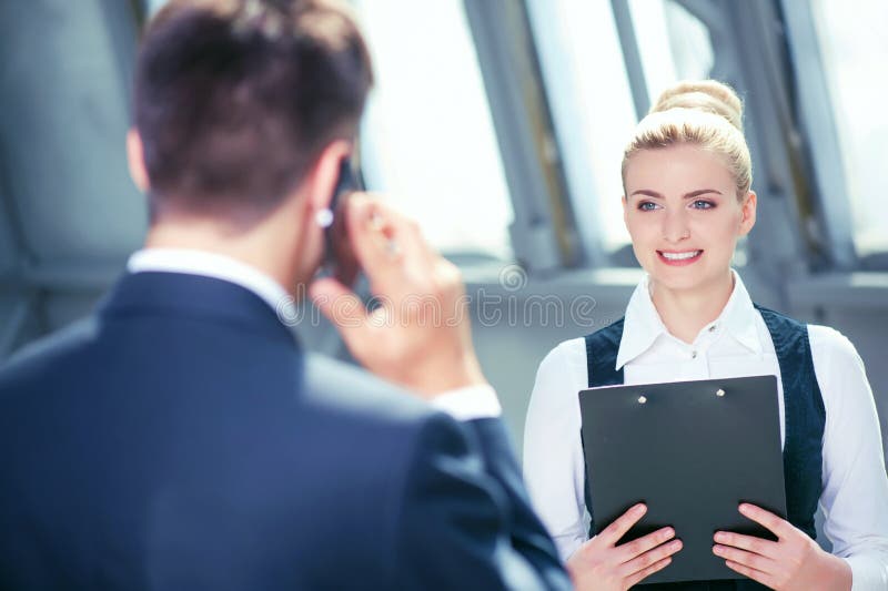 Confident businessman is talking on the phone with his client. He is looking at his female assistant. The women is holding a folder of documents and smiling. Confident businessman is talking on the phone with his client. He is looking at his female assistant. The women is holding a folder of documents and smiling