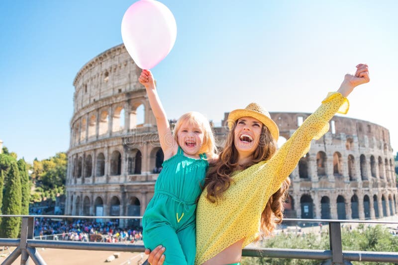 Jubiloso sonriente morena madre agotador un sombrero en verano en Roma es un posesión su rubio, cual es un feliz ondulación rosa globo.