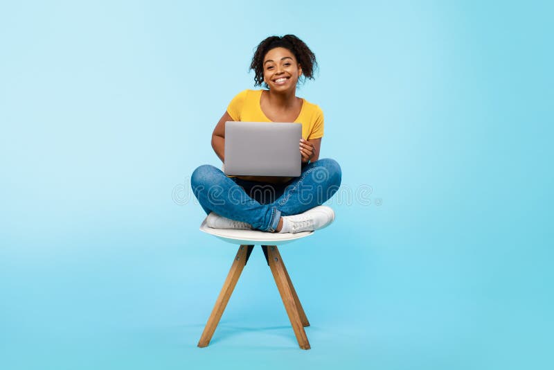 Cheery young black woman working online, sitting on chair and using laptop on blue studio background, full length stock photo