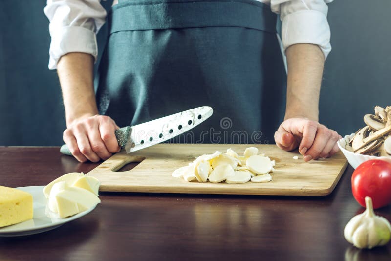 The chef in black apron cuts with a knife mozzarella cheese for pizza. Concept of eco-friendly products for cooking. Green, caucasian.