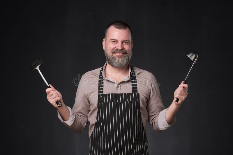 Chef europ?en m?r avec la barbe tenant une position de poche sur le mur noir de studio image stock Chef europ?en m?r avec la barbe tenant une position de poche sur le mur noir de studio image stock