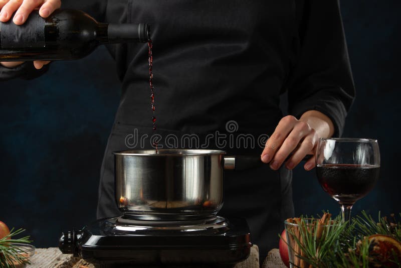 Chef pours red wine into pan for preparing mulled wine on rustic wooden table with festive composition background. Backstage of