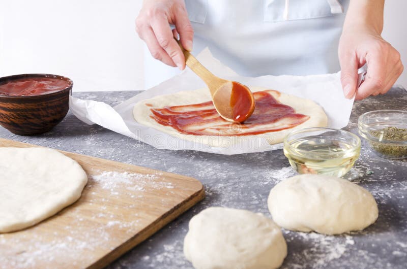 Chef préparant la pizza fraîche et répandant la sauce tomate sur la pâte Boules de la pâte, cuvettes avec de l'huile, sauce tomat photo stock Chef préparant la pizza fraîche et répandant la sauce tomate sur la pâte Boules de la pâte, cuvettes avec de l'huile, sauce tomat photo stock