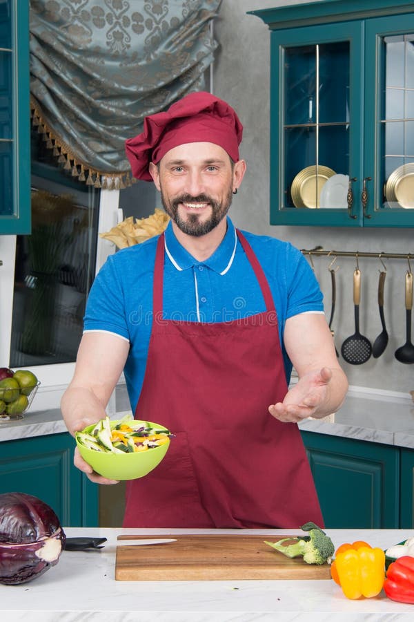 Chef wellcomes with salad. Guy in hat holds green plate with salad in hands on kitchen. stock photo Chef wellcomes with salad. Guy in hat holds green plate with salad in hands on kitchen. stock photo