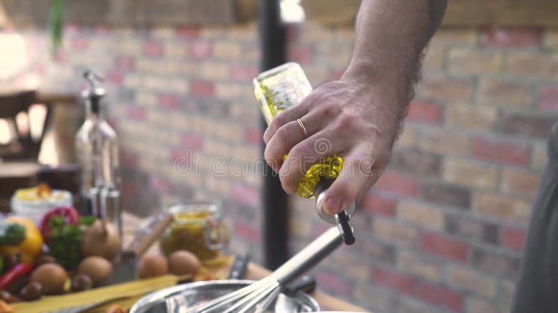 Chefkoch, der Olivenöl von der Flasche beim Kochen des Salats am Ziegelsteinhintergrund gießt Männliche Hand, die Olivenölflasche