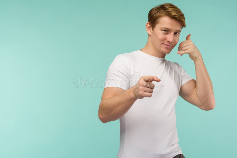 Chiamami Un giovane uomo sorridente che guarda la telecamera e gestisce il cellulare vicino alla faccia mentre sta sullo sfondo b fotografia stock libera da diritti Chiamami Un giovane uomo sorridente che guarda la telecamera e gestisce il cellulare vicino alla faccia mentre sta sullo sfondo b fotografia stock libera da diritti