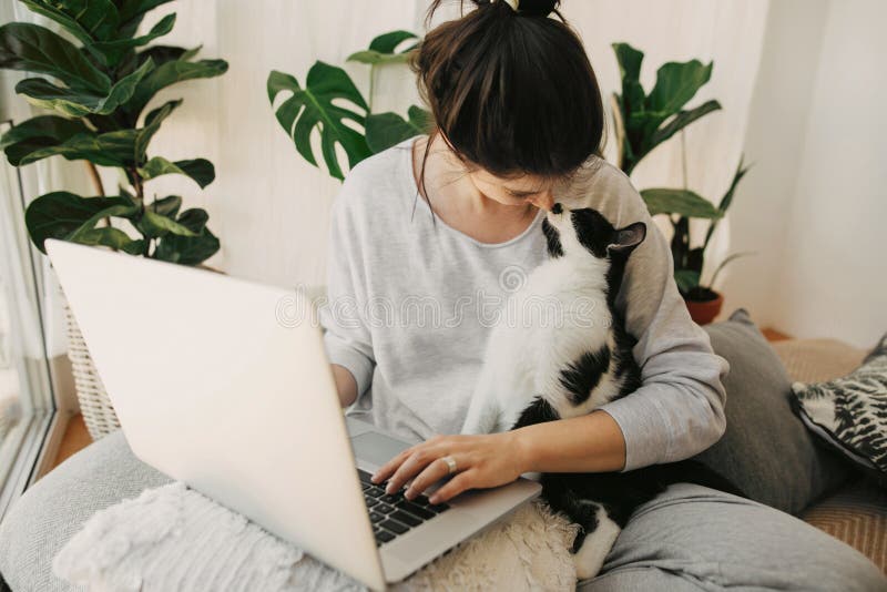 Casual girl kissing with her cat and working on laptop, sitting together in modern room with pillows and plants. Cute moments of friendship and happiness. Home office. Cat love. Casual girl kissing with her cat and working on laptop, sitting together in modern room with pillows and plants. Cute moments of friendship and happiness. Home office. Cat love