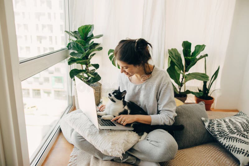 Casual girl working on laptop with her cat, sitting together in modern room with pillows and plants. Home office. Cute cat helping owner during quarantine, loyal companion. Stay home stay safe. Casual girl working on laptop with her cat, sitting together in modern room with pillows and plants. Home office. Cute cat helping owner during quarantine, loyal companion. Stay home stay safe