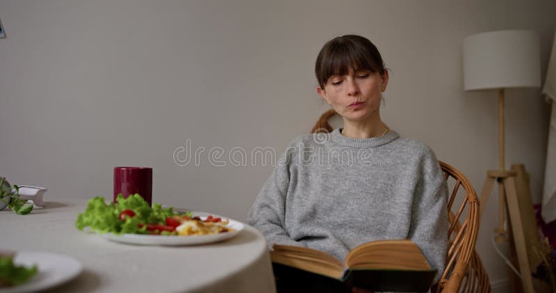 Chica con suéter gris sentada en una silla leyendo un libro y desayunando temprano en la cocina en casa metrajes