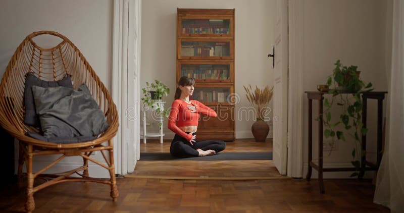 Chica con suéter naranja haciendo yoga y meditación y sentada en una pose fácil sobre una alfombra negra en el gimnasio en casa. Y metrajes