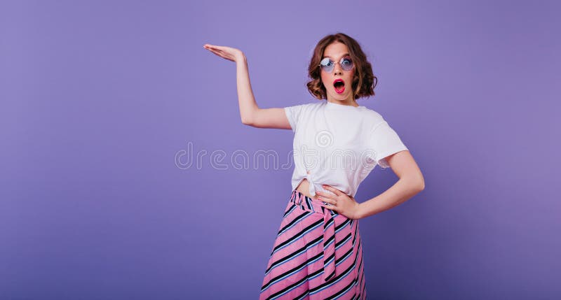 Chica elegante con cabello marrón ondulado que expresa emociones sorprendentes durante la sesión fotográfica. retrato interior foto de archivo libre de regalías