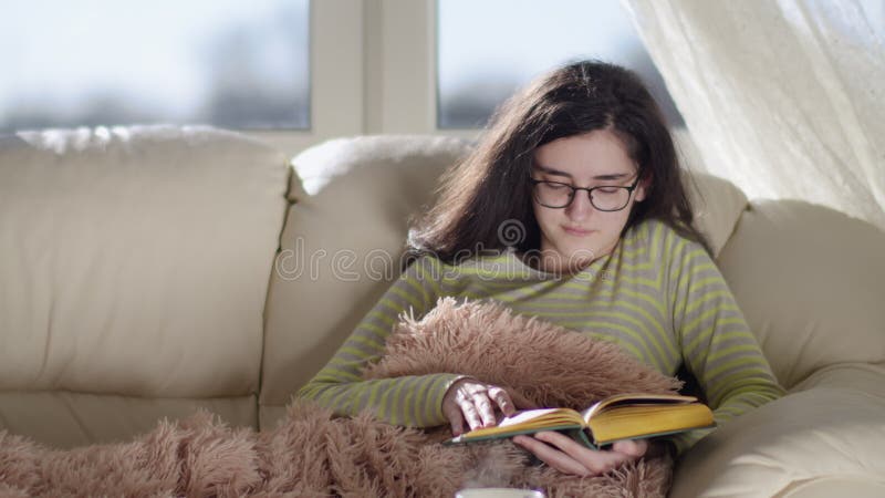 Chica feliz con anteojos tendidos en el sofá de casa leyendo un libro y sonriendo