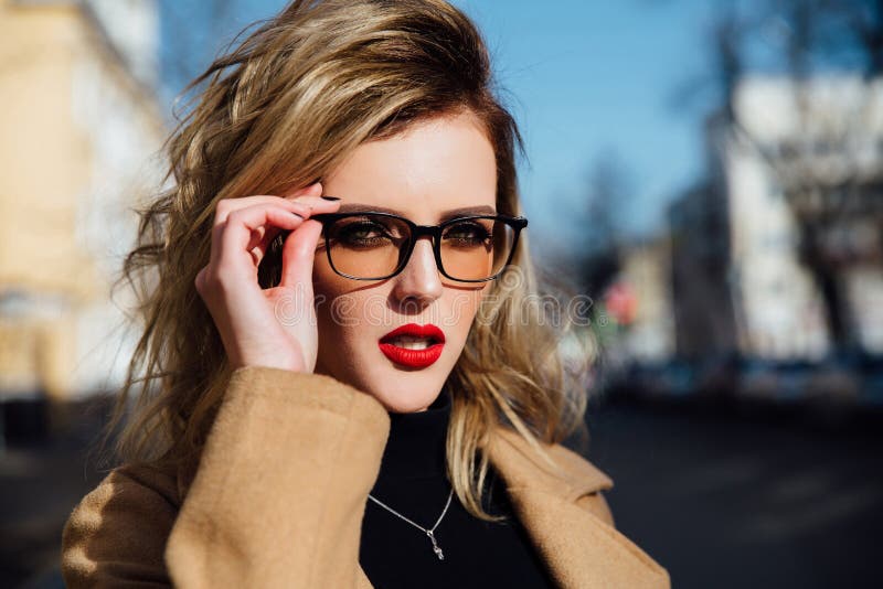 Chica joven de la moda en vidrios Labios rubios, rojos, capa beige que camina a lo largo de la calle de la ciudad imágenes de archivo libres de regalías Chica joven de la moda en vidrios Labios rubios, rojos, capa beige que camina a lo largo de la calle de la ciudad imágenes de archivo libres de regalías