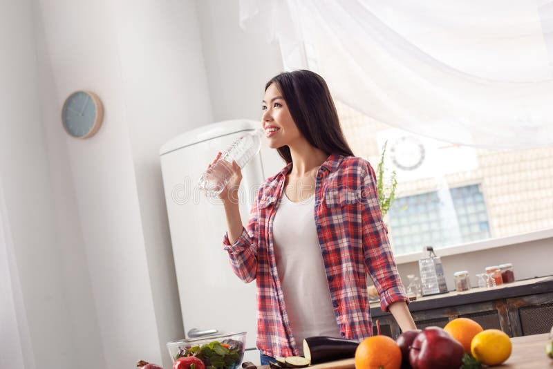 Chica joven en el agua potable permanente de la forma de vida sana de la cocina mientras que corta las verduras alegres imagen de archivo libre de regalías Chica joven en el agua potable permanente de la forma de vida sana de la cocina mientras que corta las verduras alegres imagen de archivo libre de regalías