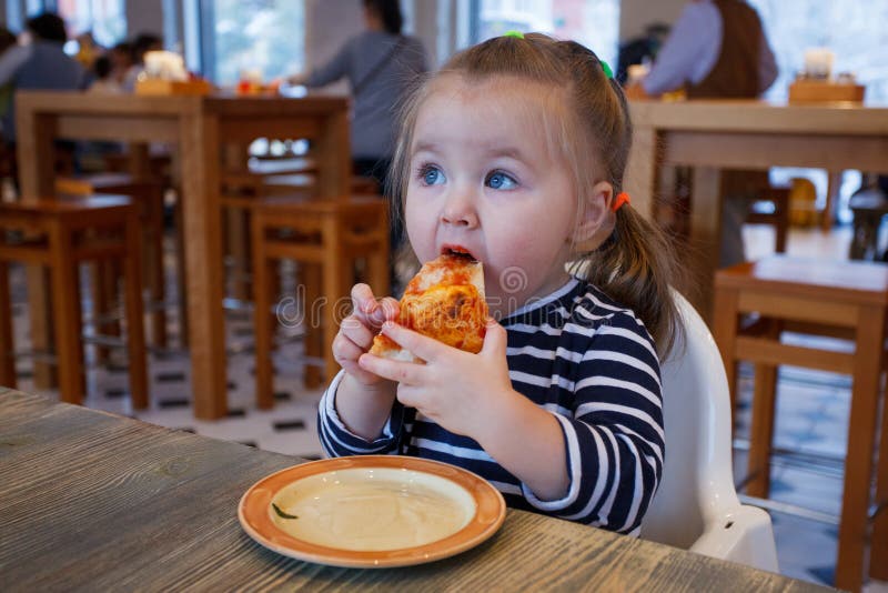 Chica joven feliz hermosa que muerde de rebanada grande de pizza hecha fresca Ella se sienta en la silla blanca en café y disfrut foto de archivo Chica joven feliz hermosa que muerde de rebanada grande de pizza hecha fresca Ella se sienta en la silla blanca en café y disfrut foto de archivo