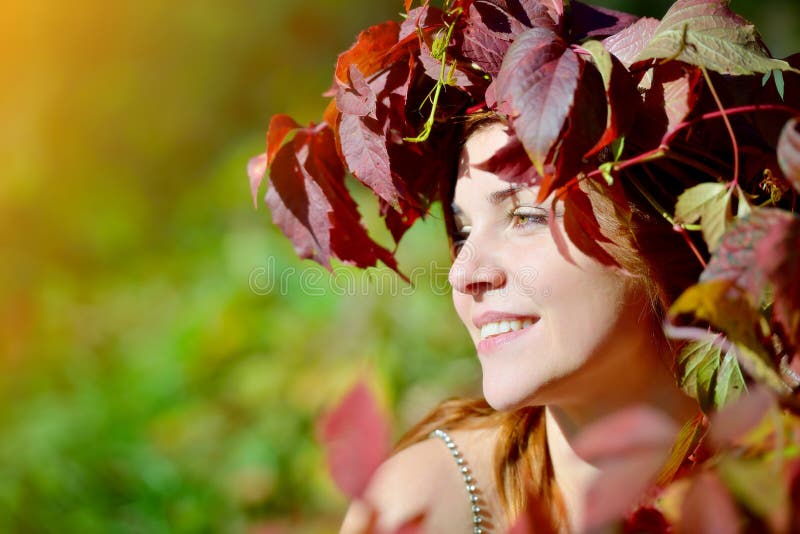 Chica joven hermosa que lleva una guirnalda de las hojas de otoño rojas y verdes que sonríen y que miran al lado en un fondo verd imágenes de archivo libres de regalías Chica joven hermosa que lleva una guirnalda de las hojas de otoño rojas y verdes que sonríen y que miran al lado en un fondo verd imágenes de archivo libres de regalías