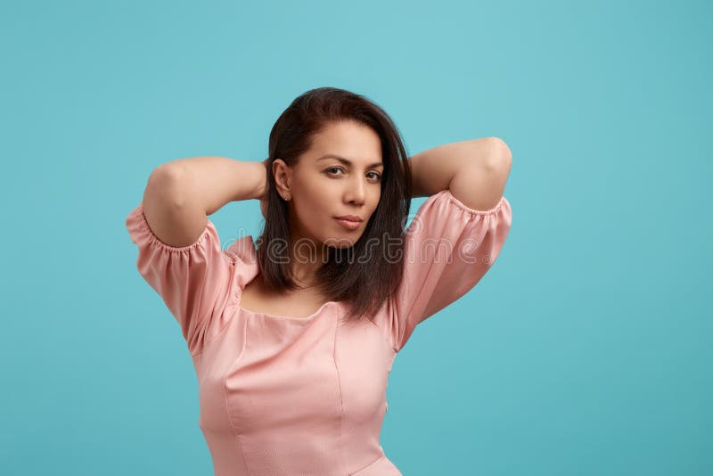 Playing girl. Portrait of beautiful young female in pink dress, keeps hands behind head, relaxed and with certainty looking at camera, isolated over blue background. People expression concept. Playing girl. Portrait of beautiful young female in pink dress, keeps hands behind head, relaxed and with certainty looking at camera, isolated over blue background. People expression concept