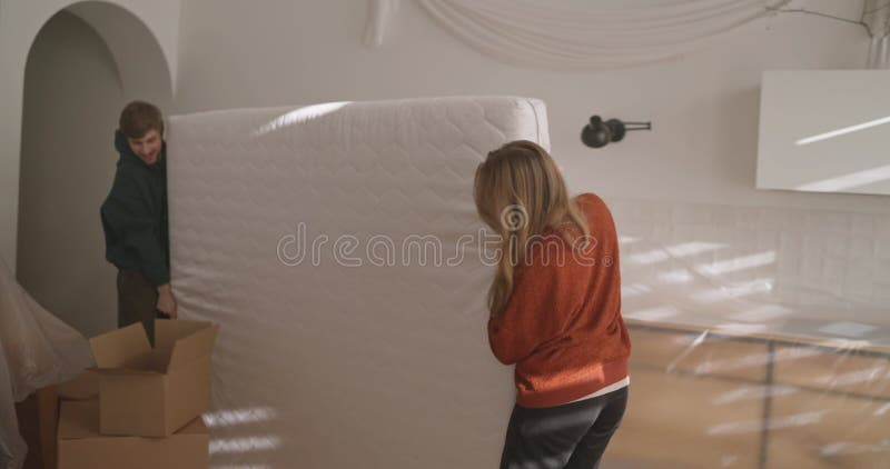Chica rubia feliz con un jersey naranja junto con su novio saca un gran colchón blanco de la habitación mientras metrajes