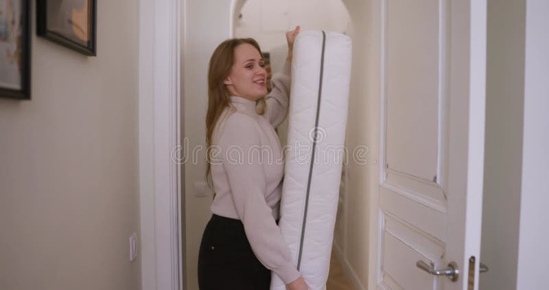 Chica rubia feliz con un suéter y su novio trae un colchón blanco a una habitación nueva mientras se muda a un apartamento nuevo metrajes
