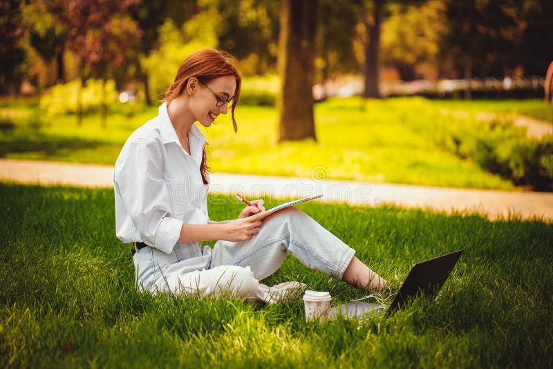 Chica sentada en el césped en el parque trabajando en la laptop pc haciendo algunas notas. profesional independiente trabajando al imágenes de archivo libres de regalías